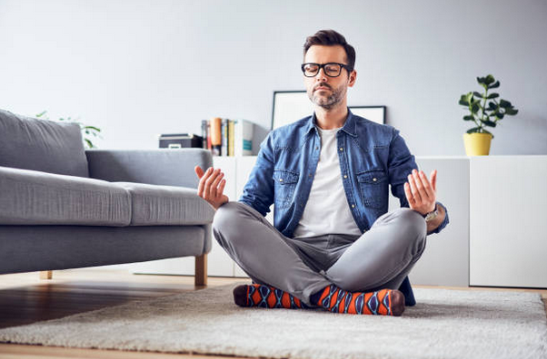 Homme assis en tailleur sur le tapis de son salon, les yeux fermés et l'air détendu